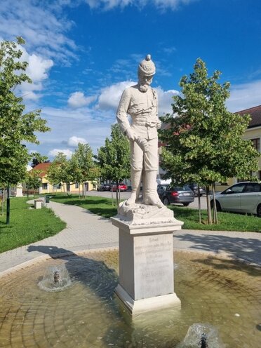 Am Bild ist eine weiße Statue eines Feuerwehrmannes zu sehen, im Hintergrund eine Straße die mit Bäumen gesäumt ist.