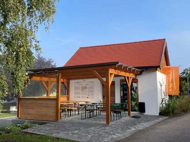 Genussrastplatz der Familie Fröschl in Staatz. Eine Holzpergola mit Heurigentischen darunter. An der Presshauswand hängt eine Übersichtstafel über das Weinviertel sowie eine Weinviertel Fahne.