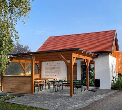 Genussrastplatz der Familie Fröschl in Staatz. Eine Holzpergola mit Heurigentischen darunter. An der Presshauswand hängt eine Übersichtstafel über das Weinviertel sowie eine Weinviertel Fahne.