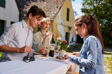 In einer Kellergasse sitzen drei Personen auf einem Tisch und trinken Wein. Im Hingergrund ist eine Kellergasse zu sehen.