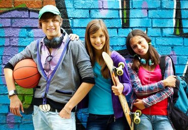 Drei Jugendliche stehen nebeneinander. Ein Bursche mit Baskertballkappe und Basketball in der Hand, ein Mädchen mit langen Haaren und einem Skatboard in der Hand sowie ein Mädchen mit Zöpfen das Köpfhörer um den Hals trägt und einen Rucksack lässig über die Schulter. Im Hintergrund ist ein gespraytes Graphity zu sehen.