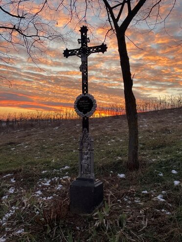 am Bild ist ein Metallkreuz Marterl mit Sonnenuntergangshimmel zu sehen.