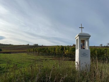 Am Bild ist ein weißes Bildstockmarterl in der Landschaft zu sehen, rundherum Felder mit blauem Himmel.